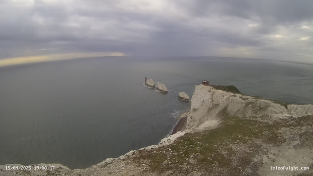 A coastal view featuring white chalk cliffs under a cloudy sky. In the distance, two lighthouses can be seen on rocky outcrops in the water, with the ocean stretching out towards the horizon. The foreground includes a grassy area with scattered stones. The overall atmosphere is calm and slightly overcast.