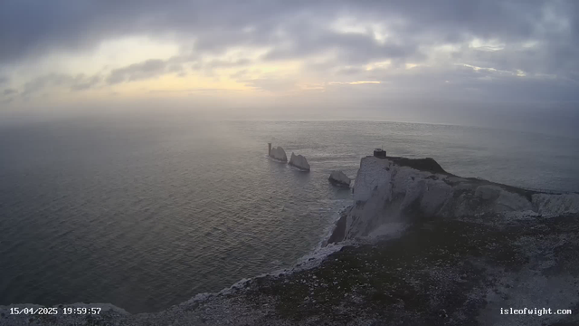 A view of a coastal landscape during twilight. The sea is calm, reflecting soft shades of gray and blue, and the sky is overcast with clouds. In the foreground, there is a cliff with a rocky surface and some vegetation. Several large white rock formations emerge from the water in the distance. A small structure is visible on top of the cliff, and a portion of a railing can be seen along the edge, suggesting a viewing area. The overall atmosphere is serene and slightly foggy.