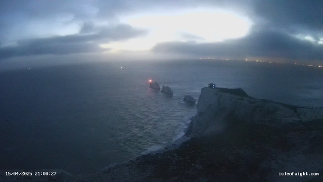 A dark, cloudy sky is visible over the sea, with waves gently lapping at the shore. In the distance, there are two rocky outcrops partially submerged in the water, one of which has a warning light glowing red. To the right, there is a white cliff edge with a structure at the top, suggesting a viewpoint or lookout. The scene is dim, indicating twilight or early evening, with some lights visible far off in the background along the horizon.