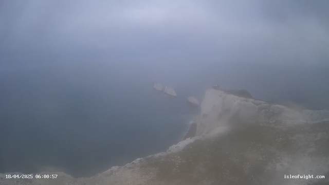 A foggy coastal scene where the horizon blends into the overcast sky. White cliffs are partially visible on the right side, leading down to a choppy, dark blue ocean. In the distance, two large rocks emerge from the water. The overall atmosphere is moody and obscured by mist. The timestamp at the bottom indicates the image was taken on April 18, 2025, at 6:00:57 AM.