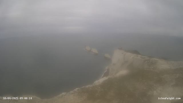 A cloudy sky above a coastal scene featuring rugged cliffs on the right side. The sea is visible in the background with some indistinct sailing vessels appearing in the water. The overall atmosphere is misty, giving the scene a soft, blurred appearance. The date and time are displayed in the lower left corner.