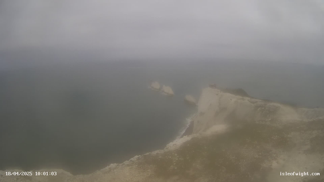 A coastal scene featuring a cliffside with grassy terrain at the forefront. In the distance, there are three visible rock formations in the water, and a small building is perched on the edge of the cliff. The sky is overcast with a grey haze, and the ocean appears calm. The image shows a sense of openness, but visibility is limited due to the cloudy weather.