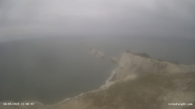 A hazy view of a coastal cliff with a rocky shoreline. The ocean is visible in the background, with several indistinct ships appearing on the water. The sky is overcast, creating a muted light in the scene. The cliff is predominantly light-colored with patches of grass or vegetation. The date and time are displayed at the bottom left corner.