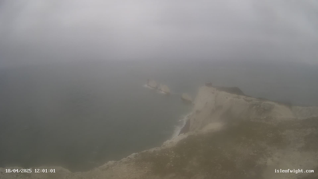 A foggy coastal scene with a rocky cliff on the right side, overlooking a dark, choppy sea. The sky is overcast and the visibility is low. In the distance, several large rocks emerge from the water. There is a small structure at the top of the cliff. The overall atmosphere is gray and misty, suggesting a chilly and damp environment.