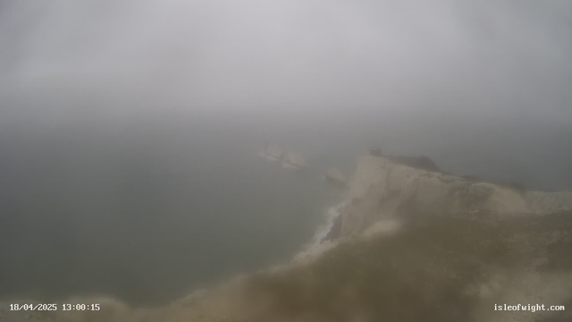 A foggy coastal scene with a rocky shoreline. The sky is overcast, blending gray clouds with the horizon. The water appears dark and choppy, and faint shapes of rocks are visible in the water. The image has a muted color palette due to the fog. In the distance, a slight outline of land and a structure can be seen at the edge of the cliffs. The date and time are displayed in the bottom left corner.
