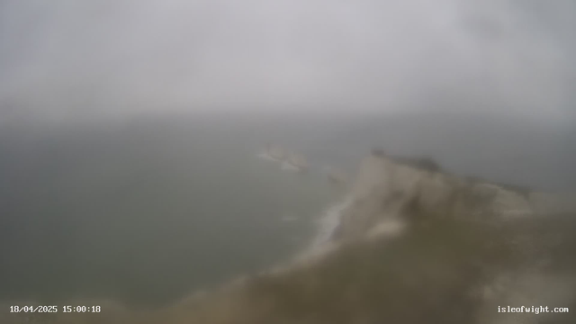 A foggy coastal scene with a blurred view of the sea and cliffs. The image is overcast, making visibility poor, with indistinct shapes of landforms in the distance. The foreground shows a rocky edge leading to the water. The timestamp indicates the image was taken on April 18, 2025, at 15:00:18.