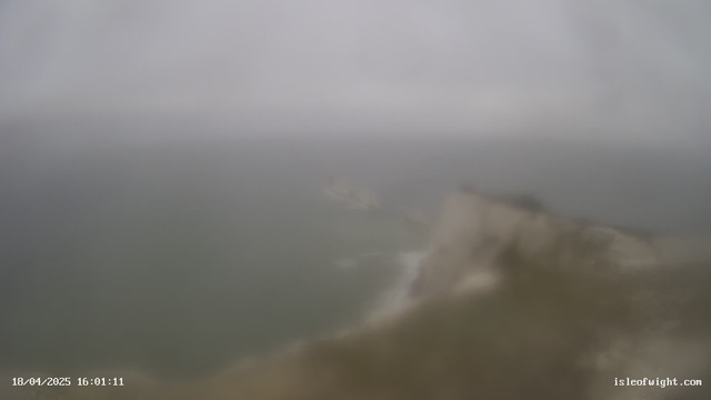 A foggy coastal scene overcast with gray clouds. The ocean appears choppy with low waves, and a rocky shoreline can be seen in the distance. The image is blurred, creating a misty atmosphere with limited visibility. The date and time in the corner indicate April 18, 2025, at 16:01:11.
