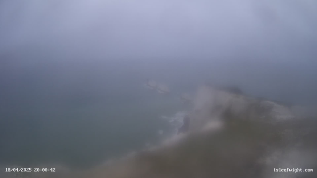 A foggy scene over the ocean, with indistinct shapes of land and possibly boats visible through the mist. The colors are muted and primarily gray and blue, creating a subdued atmosphere. The view is slightly blurry, suggesting poor visibility, likely due to weather conditions. A timestamp at the bottom indicates the date and time of the image.