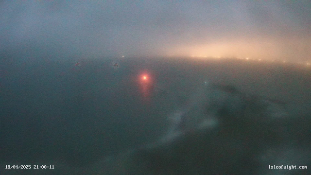 A foggy view of the ocean at night. A faint red light is visible on the water's surface, possibly from a buoy or lighthouse. The background is obscured by mist, with soft, diffused lights appearing in the distance, suggesting the presence of land or buildings. The overall color scheme is dark and muted, with various shades of gray and hints of warmth from the lights. The date and time are displayed in the lower left corner.