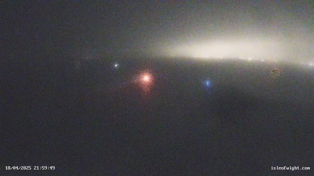 A foggy scene with dim visibility. A large area of greyish fog fills the frame, with faint, colorful light spots visible in the distance. The lights include a bright red light in the center, a blue light to the right, and a white light further away. The background shows a soft glow, hinting at lights from buildings or vehicles obscured by the fog. The date and time are displayed in the bottom left corner.