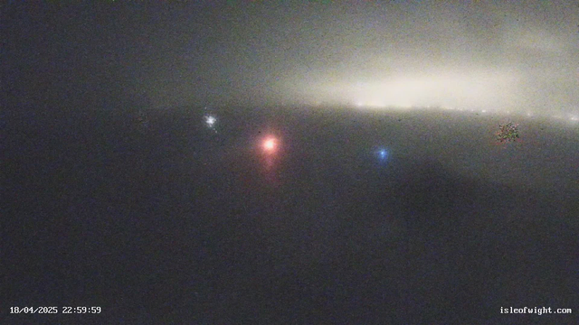 The image shows a foggy and dark scene, with faint lights in the distance. There are two small colored lights: one red and one blue, slightly visible against a backdrop of hazy darkness. The overall atmosphere is misty, giving a sense of obscured visibility. There are also dim lights scattered across the horizon, suggesting distant buildings or vehicles.
