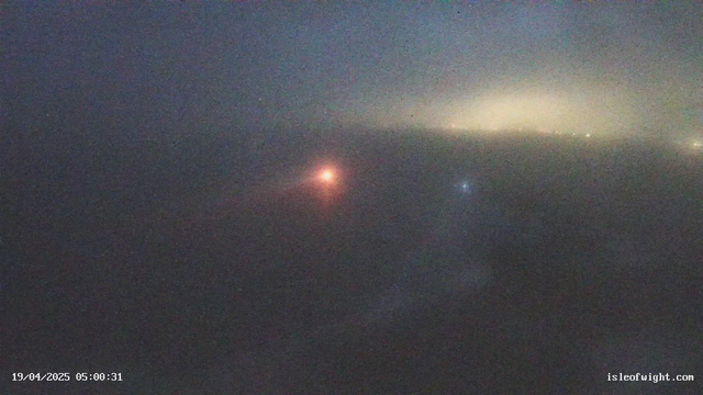 A foggy scene captured by a webcam, where visibility is low. Two lights are partially visible: one is a bright orange to the left, and the other is a softer blue to the right. The background is mostly obscured by fog, giving a muted overall appearance. The date and time in the lower left corner reads "19/04/2025 05:00:31."
