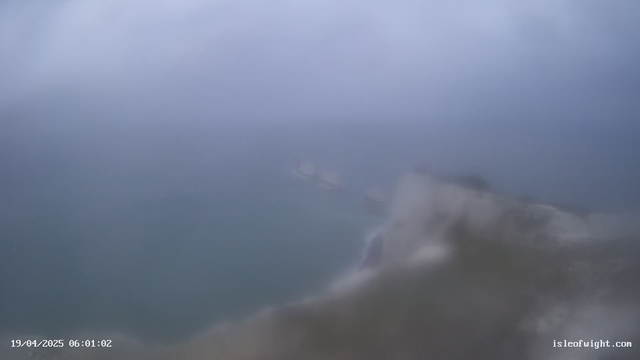 A foggy and blurred coastal scene featuring a turbulent sea under a cloudy sky. The visibility is low, and indistinct shapes of rocks or land can be faintly seen in the background. The date and time are displayed at the bottom left corner of the image.