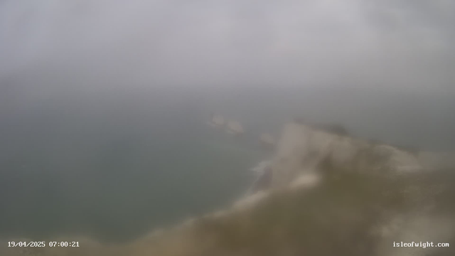 A blurry image showing a coastal view during foggy weather. The sea is visible, with a gray sky and indistinct shapes of rocks or cliffs along the shoreline. Visibility is low, creating a hazy atmosphere. The timestamp at the bottom reads April 19, 2025, at 07:00:21.
