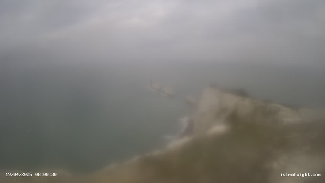 A blurred webcam image depicting a coastal scene under overcast skies. The ocean is visible in muted colors, with fog or mist obscuring the view. The shoreline is partially visible, along with some indistinct structures or rocks in the water. The image appears hazy and lacks sharp detail, suggesting dreary weather conditions. A timestamp at the bottom indicates the date and time.