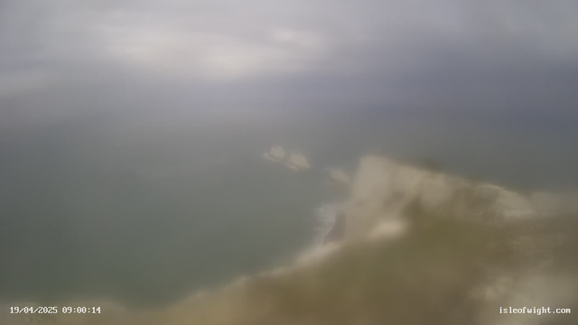A cloudy sky casts a dull light over a calm sea. In the distance, a faint outline of rocky formations can be seen above the water, while the foreground is mostly blurred and indistinguishable. The shoreline appears to have a sandy texture, merging into the subtle waves of the water.