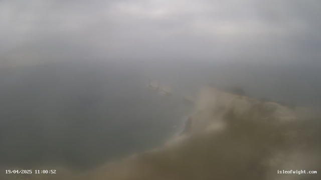 A cloudy sky with limited visibility over the ocean. The water appears choppy, and there are blurred outlines of coastal rocks or cliffs in the foreground. A faint silhouette of a structure is visible on the water's surface. The overall scene conveys an overcast and somewhat misty atmosphere.