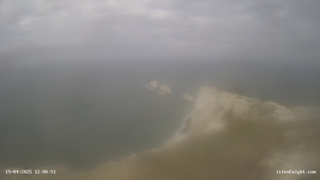 A coastal view with cliffs meeting the ocean, partially obscured by mist. The sky is overcast with clouds, and the water appears calm and gray. Some rocky formations are visible just off the shore. The image has a hazy quality, suggesting a lack of visibility.