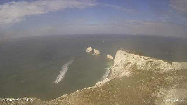 A coastal scene depicting rocky cliffs under a blue sky with scattered clouds. In the foreground, rugged white cliffs slope down toward the sea. A motorboat is visible on the water, leaving a white wake as it moves. In the distance, three prominent rock formations are partially submerged in the ocean. The water appears calm with some gentle ripples.