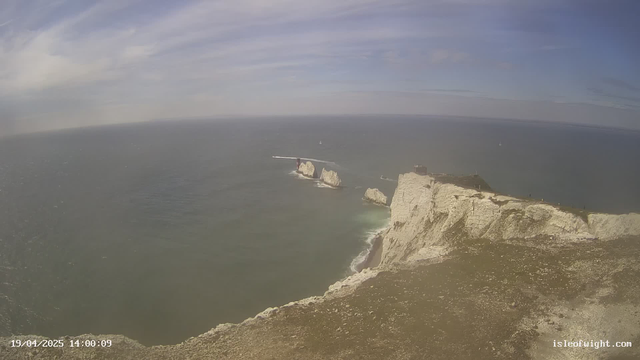 A coastal scene shows a vast expanse of blue ocean stretching to the horizon under a partly cloudy sky. In the foreground, rocky cliffs with white chalk formations edge the water. A small boat is visible near the cliffs, and two larger white rock formations protrude from the ocean. Several people can be seen near the edge of the cliffs. The overall atmosphere is serene and coastal.
