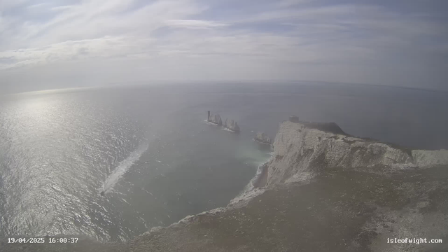 A panoramic view of a coastline with cliffs on the right side. In the distance, several rock formations emerge from the ocean, some of which are tall and jagged. The water is shimmering with the reflection of sunlight, and the sky is mostly cloudy with light gray and blue hues. A narrow boat is visible, creating a wake as it moves through the water. The scene conveys a sense of tranquility and natural beauty.