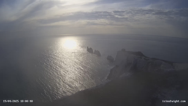 A coastal scene featuring calm, reflective waters extending towards the horizon under a cloudy sky. The sun is positioned low in the sky, creating a bright reflection on the water surface. In the foreground, rugged cliffs and rock formations jut out from the land, with a small structure visible on one of the cliffs. The overall atmosphere is serene and slightly overcast, suggesting a tranquil moment at dusk.