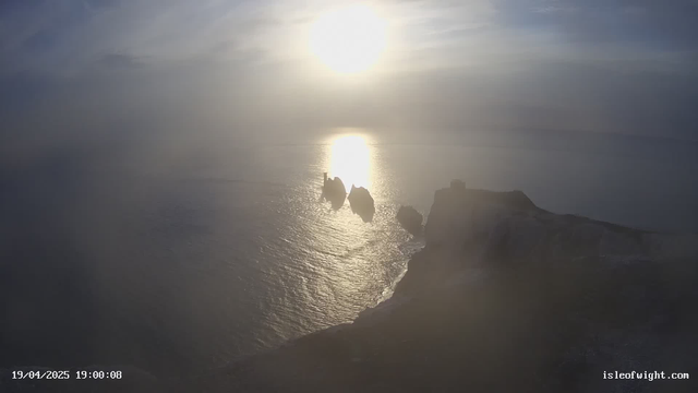 A landscape view of a coastal area during sunset, with a bright sun low in the sky reflecting on the water. Silhouetted against the light, three distinct rock formations are visible in the water, along with a steep cliff from which the view is taken. The scene is slightly shrouded in fog, creating a soft, muted atmosphere. The date and time are displayed in the lower left corner.