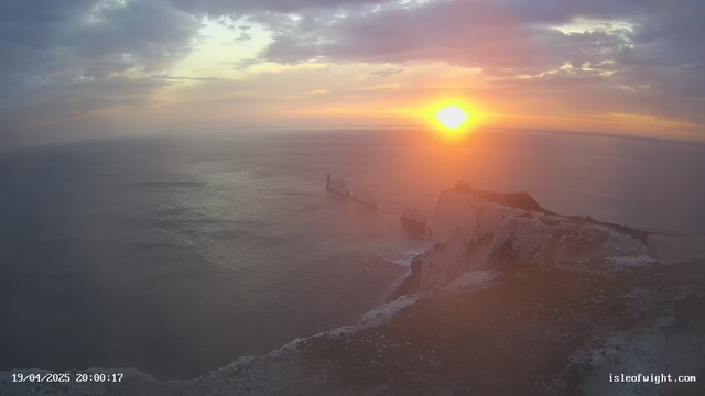 A panoramic view of the ocean during sunset, with the sun low on the horizon emitting warm orange and yellow hues. The sky is partly cloudy with soft, diffused light. In the foreground, rocky cliffs are visible, and a few rock formations emerge from the water, suggesting a rugged coastline. The sea reflects the colors of the sky, creating a serene and tranquil atmosphere.
