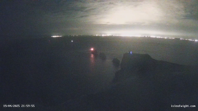 A dark scene showing a coastal view at night. On the left side, there is a faint red light from a boat or beacon reflecting on the water. The water appears calm, and there are irregular dark shapes, likely rocks or land, partially submerged. In the background, faint lights suggest the outline of a distant shoreline or town, appearing as a faint glow against the dark sky. The sky is mostly cloudy, with a slight haze, and the time and date are displayed at the bottom left corner.