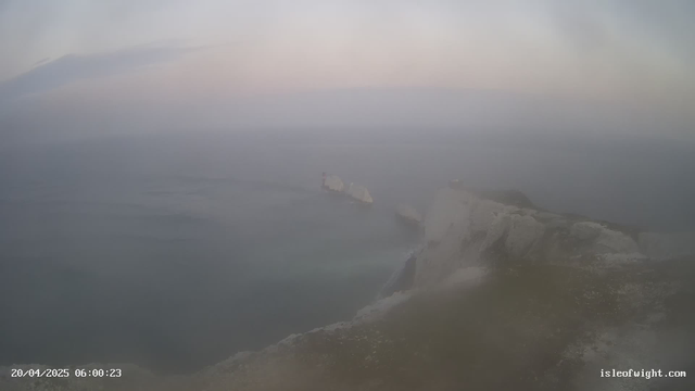 A foggy seaside scene at dawn. The sky is a pale blue with soft clouds. In the foreground, a rocky cliff edge descends to the calm ocean below. A distant landmark, possibly a lighthouse or a rock formation, is partially obscured by mist. The water reflects muted hues from the sky, creating a serene but hazy atmosphere. The bottom left corner shows a timestamp indicating the image was taken on April 20, 2025, at 6:00 AM.