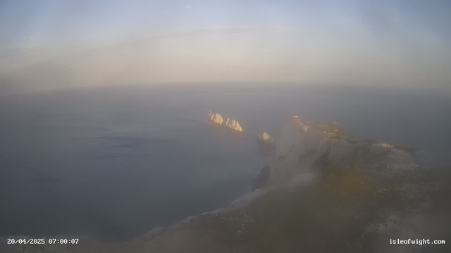 A coastal view at dawn, with soft blue and gray hues blending in the sky. The ocean appears calm, reflecting the subtle light. In the foreground, there's a rocky cliff with jagged edges leading into the water. Several large, white cliffs rise along the shoreline, partially obscured by mist. A hint of a red structure can be seen on one of the cliff faces, adding a contrasting color to the scene. The overall atmosphere feels serene and tranquil.