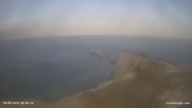 A coastal view showing a cliff edge in the foreground with a rocky shoreline. The ocean extends toward the horizon, appearing calm with subtle waves. The sky is slightly cloudy, creating a soft, hazy atmosphere. Two sea stacks can be seen in the water to the left, and the overall color palette features muted blues and greens. The timestamp below indicates the date and time.