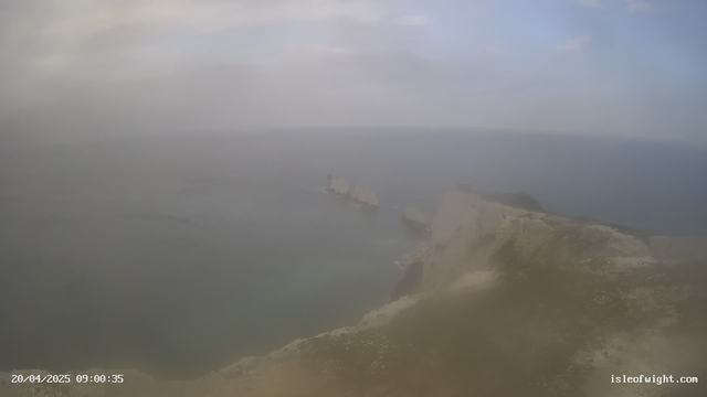 A coastal scene shows a rocky cliff on the right side of the image, gradually descending into a misty ocean. In the distance, there are several large, white rock formations protruding from the water, with a partially visible structure, likely a lighthouse, on one of them. The sky overhead is grey and overcast, contributing to a hazy atmosphere. The bottom left corner displays the date and time.