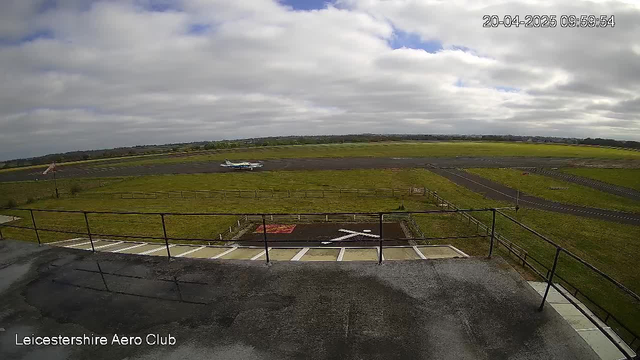 A view of an airport from a webcam positioned on a building. The foreground shows a flat rooftop with a railing, and a white cross marking on the ground. In the background, a small aircraft is taxiing on a runway surrounded by green grass. The sky is cloudy with varied shades of gray and blue. The scene indicates a calm day at the airport.