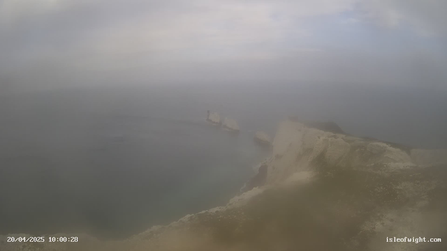 A foggy coastal scene with a view of the ocean. In the foreground, there are rocky cliffs and a few rock formations emerging from the water. The sky is overcast, and visibility appears limited, giving the image a misty appearance. The date and time are visible in the lower left corner.