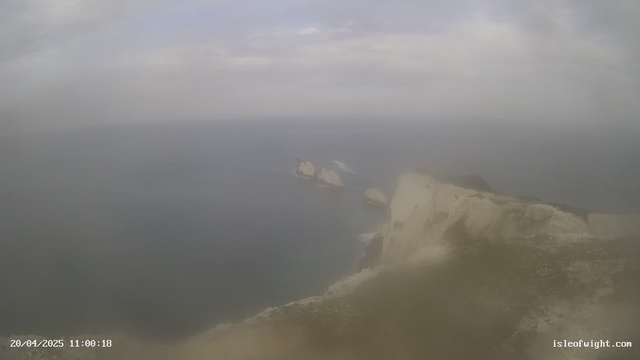 A foggy coastal scene shows the edge of a cliff over the ocean. In the distance, there are two rocky outcrops, slightly obscured by the mist. The sky is cloudy with soft gray tones, and the water appears calm, blending into the horizon where it meets the sky. The bottom of the image shows grassy cliffs, while the time and date display is in the lower-left corner.