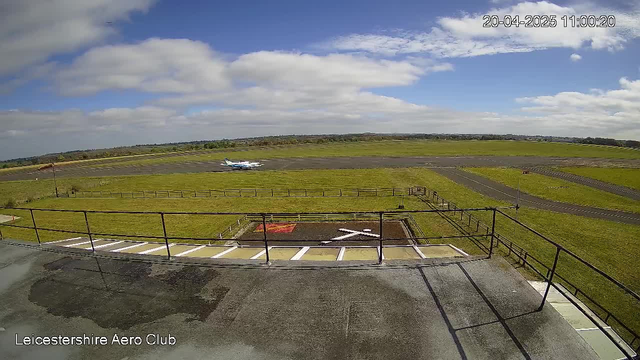 A clear view of an airstrip at the Leicestershire Aero Club, with a small plane taxiing on the runway. The landscape is predominantly green grass, with a few scattered trees in the background. The sky is bright blue with some white clouds. In the foreground, there is a paved area with a helipad marked by a red and white "X," and a railing visible along the edge of the platform.