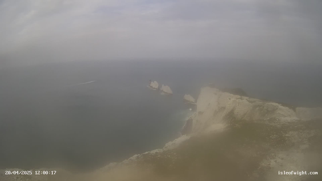 A panoramic view of a coastal landscape featuring rocky cliffs on the right side. Several white rock formations rise from the calm water in the middle ground. In the background, the sea stretches out toward an overcast sky. A small boat can be seen leaving a trail in the water, moving away from the shore. The foreground includes a grassy area with light-colored earthy tones. The image has a soft, hazy quality due to atmospheric conditions.