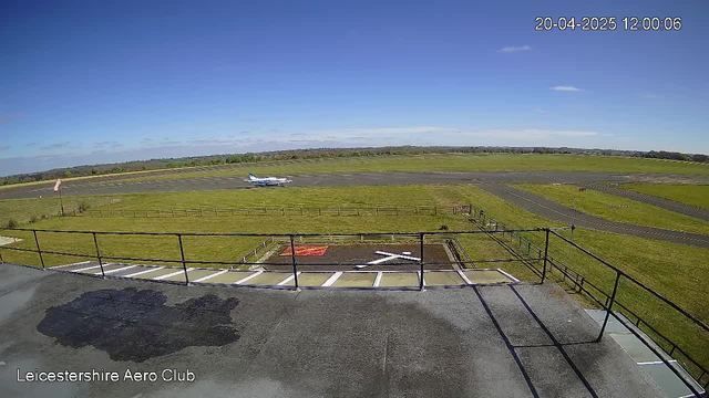 A clear blue sky with some clouds above a green field. In the foreground, there is a paved area with a white "X" marking. A small plane is seen taxiing on a runway in the distance. The overall scene appears to convey a calm and sunny day at an aerodrome.