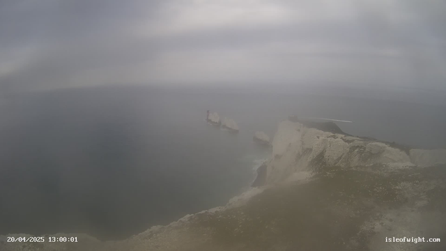 A coastal scene featuring cliffs on the right side, overlooking a gray, cloudy sea. In the distance are rocky formations emerging from the water. The sky is overcast, creating a foggy atmosphere. A small boat can be seen on the left side, leaving a trail in the water. The image depicts a sense of serenity and natural beauty despite the dull conditions.