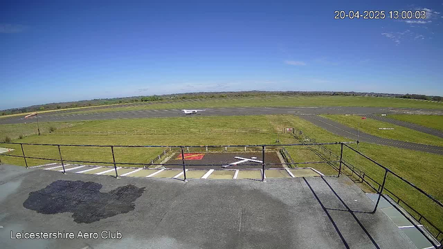 A clear blue sky with a few clouds is visible above a wide green field and an airport runway. In the foreground, there is a railing and a patch of grey surface with some dark stains. A light aircraft is seen on the runway, moving towards the left side of the image. To the right, a wind indicator is present, and there are noticeable runway markings. The overall scene gives the impression of a busy aerodrome on a sunny day.