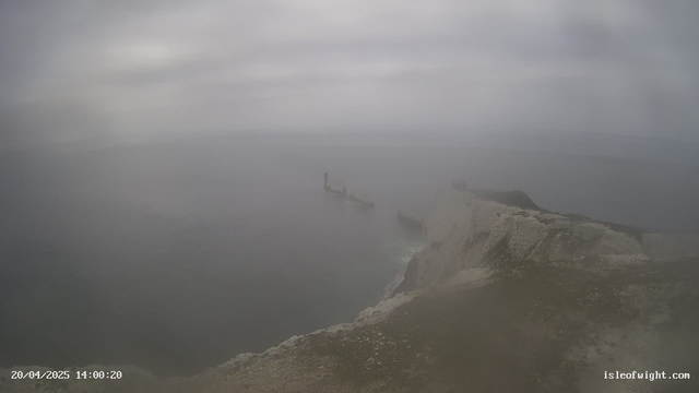 A coastal scene with a rocky shoreline and cloudy skies. In the distance, a faint outline of a shipwreck is visible on the water's surface. The ocean appears calm, with a grayish hue blending into the overcast sky. The foreground shows a grassy cliff edge leading down to the water, highlighted by patches of white rock. The overall atmosphere is misty and subdued.