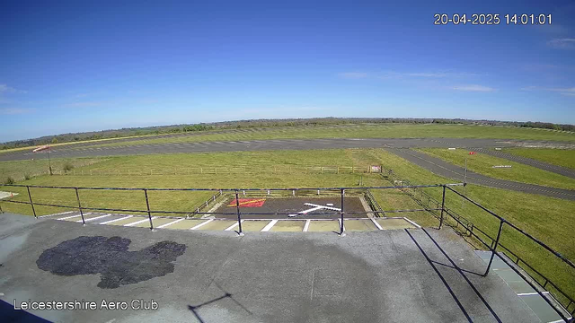 A clear blue sky is visible above a grassy landscape. In the foreground, there is a flat rooftop with a railing, overseeing a wide, open airstrip below. The airstrip is mostly paved with a few markings, including a red sign and a white cross marking, indicating a helicopter landing area. In the distance, green fields stretch out towards the horizon. The overall scene is bright and sunny, depicting a calm day at an aero club.