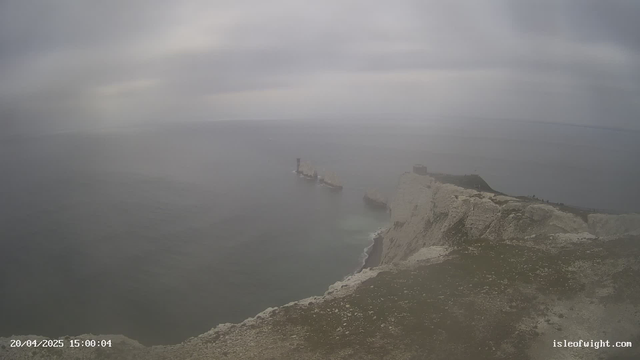 A cloudy sky looms over a coastal scene featuring steep cliffs and a rugged shoreline. In the distance, several large boats are visible near a rocky outcrop. The sea is calm, with gentle waves lapping at the base of the cliffs. The landscape is primarily gray and muted, with some patches of green grass on the cliff edges.