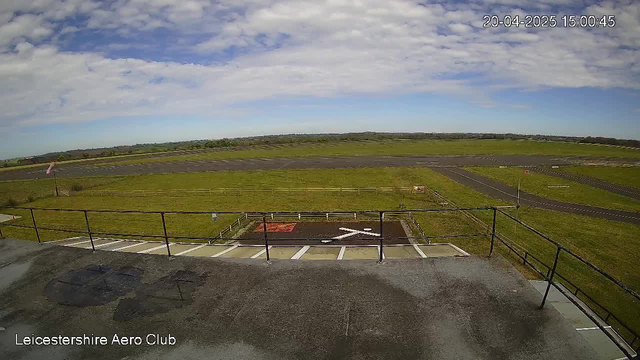 A view from a webcam at Leicestershire Aero Club overlooking an empty airfield. The foreground shows a railing and a section of pavement, while the middle ground is a grassy area with a white "X" marking on the ground, indicating a landing zone. The background features a long, straight runway bordered by a fence, with patches of grass, and a clear blue sky with some fluffy clouds.