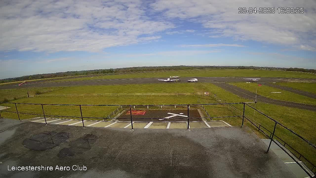 A view from a webcam at Leicestershire Aero Club shows a large green field with a runway in the background. A small airplane is located on the tarmac, and a vehicle is nearby. The sky is mostly clear with scattered clouds. In the foreground, a railing and some markings on the ground are visible, indicating a designated area, with a faint outline of a "X" shape. The image timestamp indicates it was captured in the afternoon.
