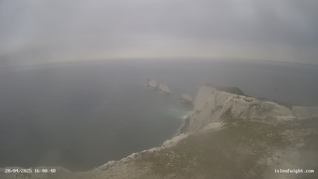 A coastal scene shows a gray, overcast sky above a calm sea. The water appears dark and reflective, with gentle waves. A prominent chalk cliff rises on the right side, where the land is grassy and uneven. In the distance, there are three rocky outcrops emerging from the water, with a lighthouse structure on one of them. The foreground features rocky terrain leading down to the shore. The overall atmosphere is misty and serene. The date and time are displayed in the bottom left corner.