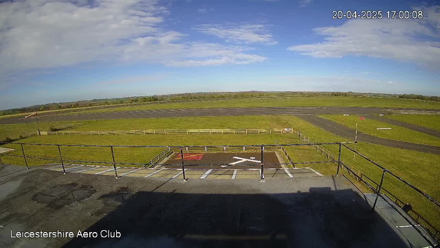 A clear sky with a few clouds is visible above a green landscape that includes an airstrip. In the foreground, there is a section of a building's roof with railings and a view of a helipad marked with a white 'X' on a dark surface. Surrounding the helipad, there are white markers and a patch of red ground. The airstrip is visible in the distance, along with a fence outlining the area. The image is bright and displays a sunny atmosphere.