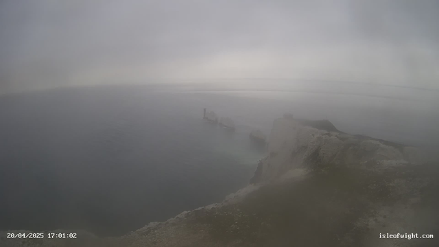 A foggy webcam view showing a cloudy sky above a calm sea. In the foreground, there is a rocky shoreline with a slight incline. Off the coast, partially obscured by mist, are the remains of a structure extending into the water, along with several large rocks. The overall scene has a muted color palette, dominated by grays and blues, creating a hazy atmosphere.