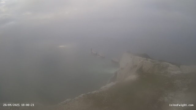 A foggy landscape view showing a rocky coastline meeting the ocean. The water is visible but appears vague due to the mist. In the distance, some rocky formations are partially obscured by the haze. The sky is overcast, creating a melancholic atmosphere. A timestamp at the bottom indicates the date and time of the image.